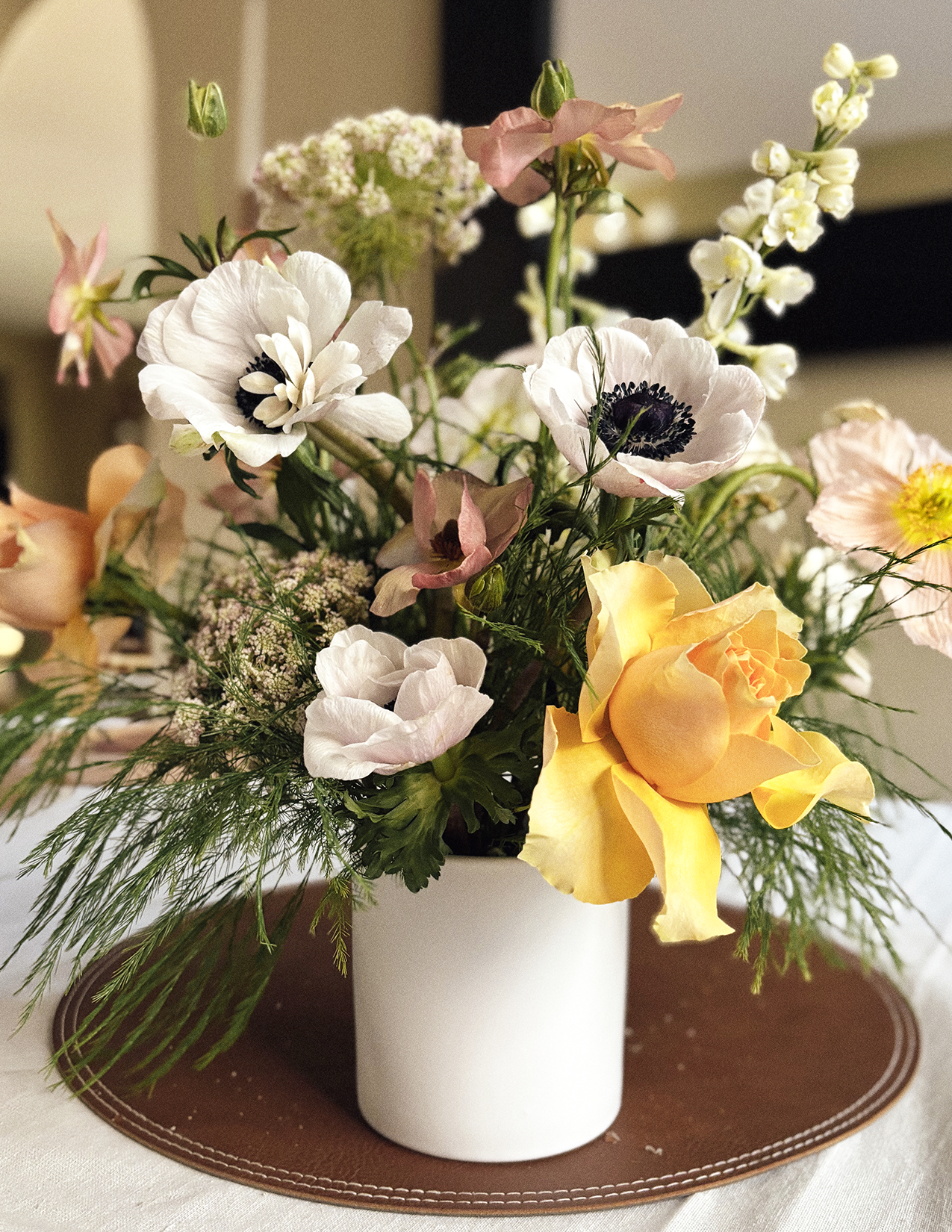 A floral arrangement in a white vase with white, pink, yellow, and purple flowers along with green foliage, placed on a brown placemat.