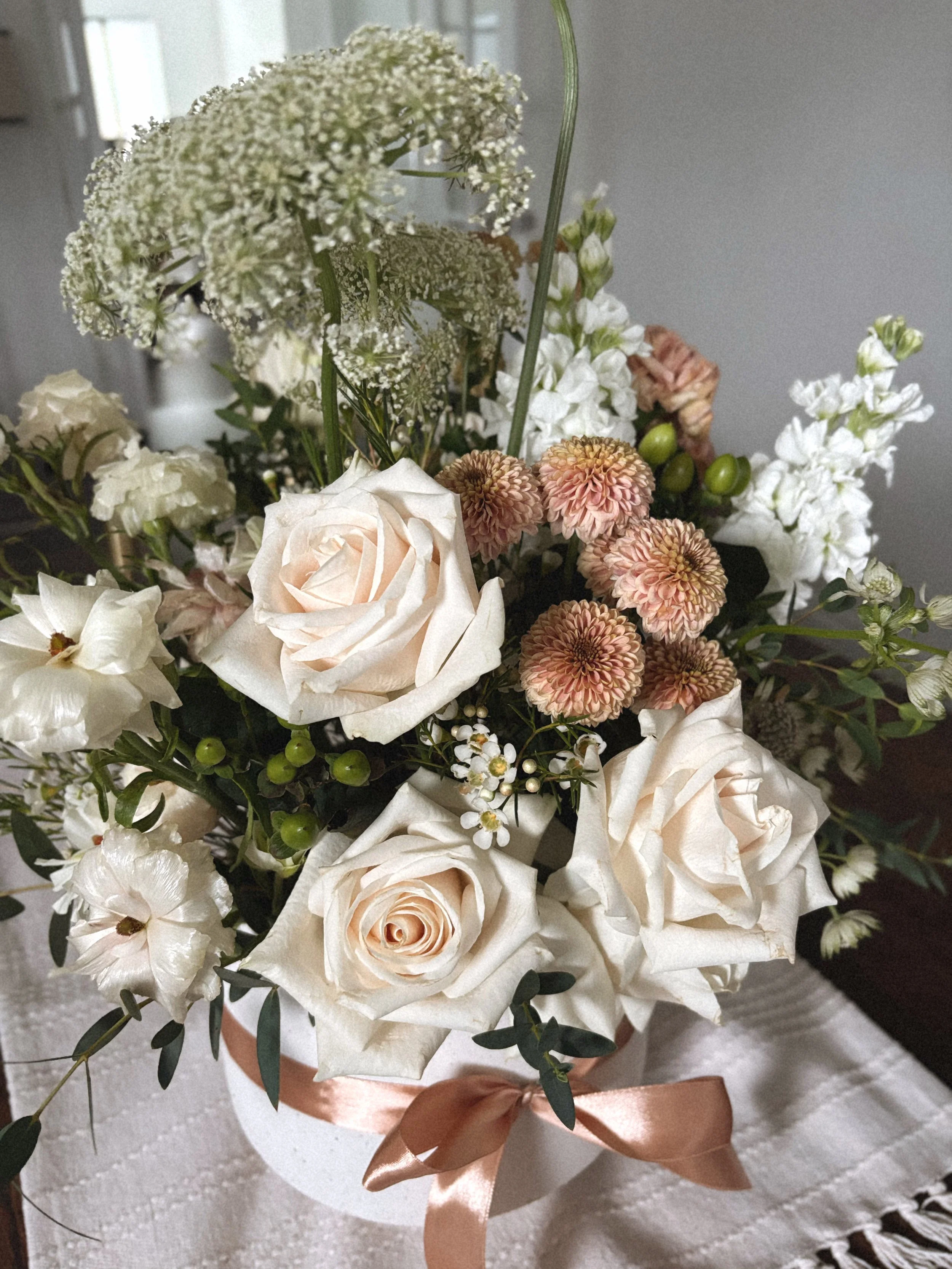 A bouquet of mixed flowers including white roses, pink chrysanthemums, white hydrangeas, and Queen Anne's lace, wrapped with a satin ribbon, placed on a white textured fabric surface.