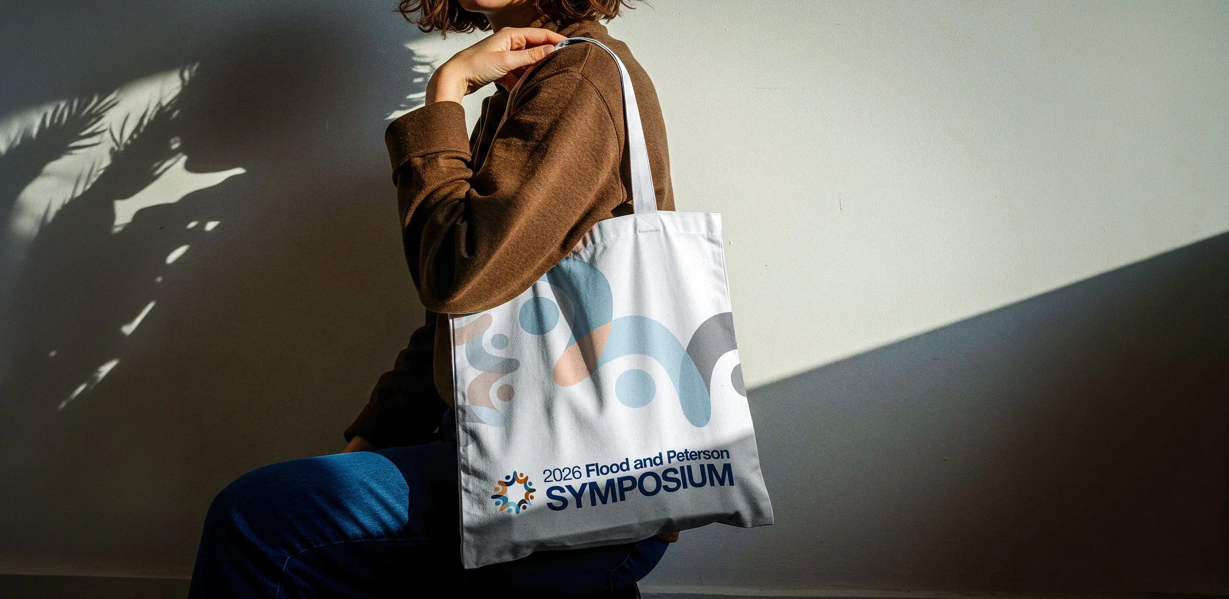 A person holding a tote bag that reads '2026 Flood and Peterson Symposium' with a colorful circular logo. The person is wearing a brown long-sleeve shirt and dark jeans, sitting against a plain wall with shadows cast on it.