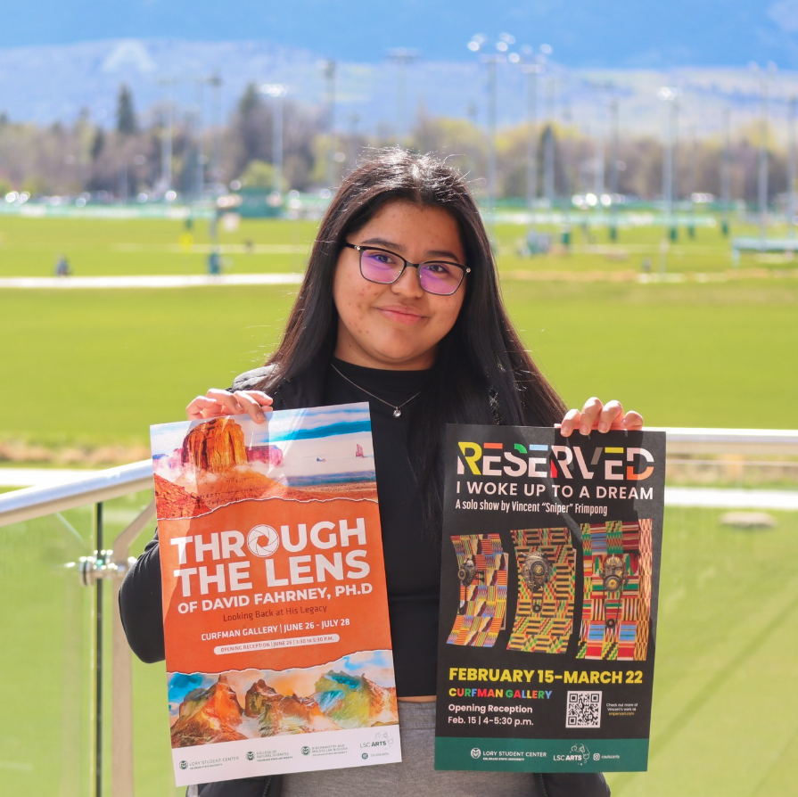 A young woman with glasses and long dark hair holding two posters at an outdoor event.