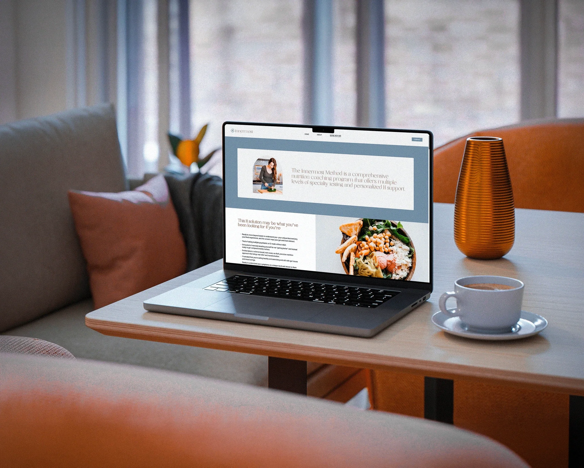 A laptop on a wooden desk displaying a health and nutrition coaching website, with a bowl of salad in front of it, a cup of coffee on a saucer, and a tall orange striped vase on the right side. In the background, there are curtains and a window.