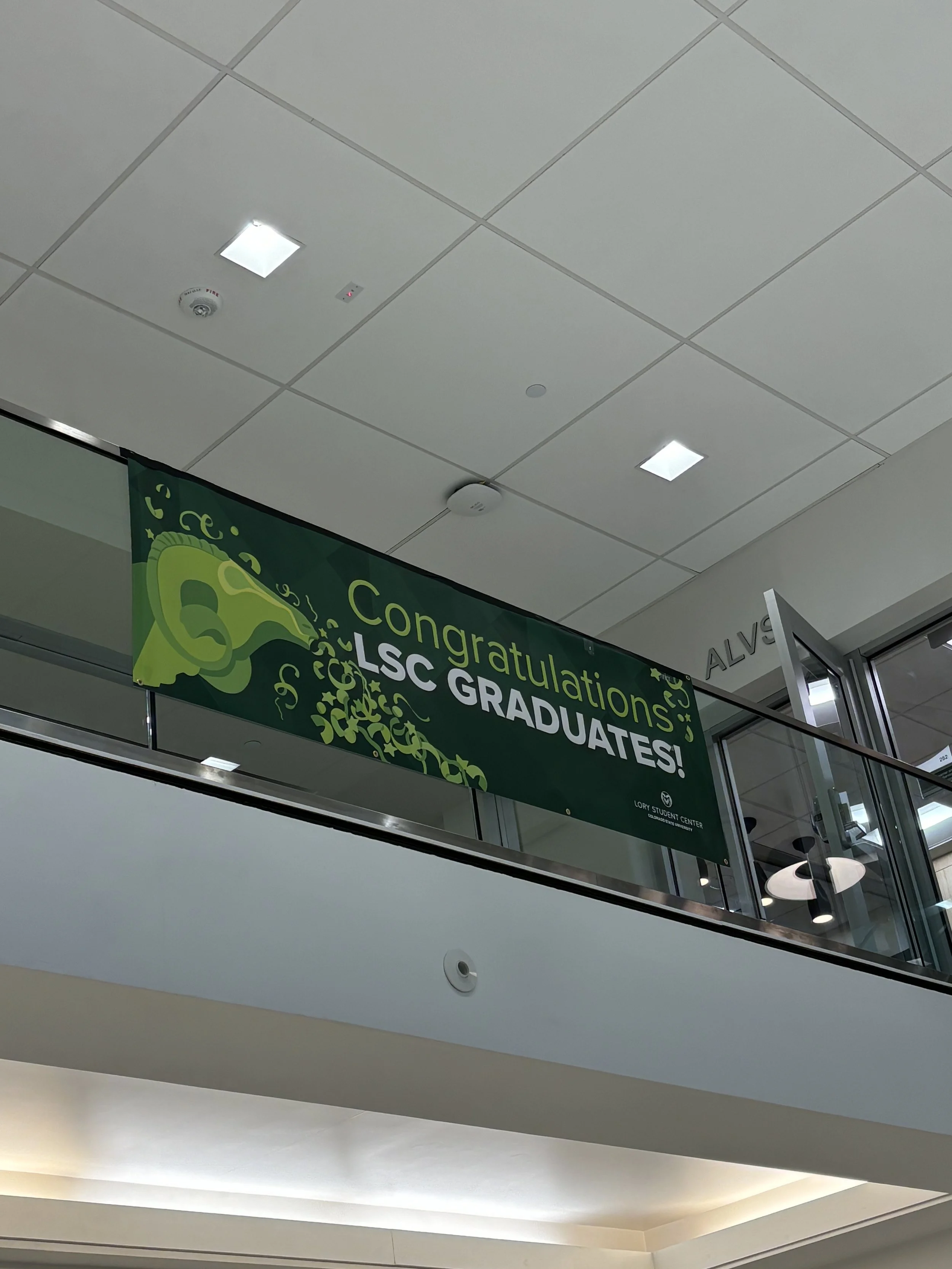 A green banner hangs in an indoor space with the text 'Congratulations LSC Graduates!' The ceiling has white tiles, with lights and fire alarm, and a glass railing is visible.