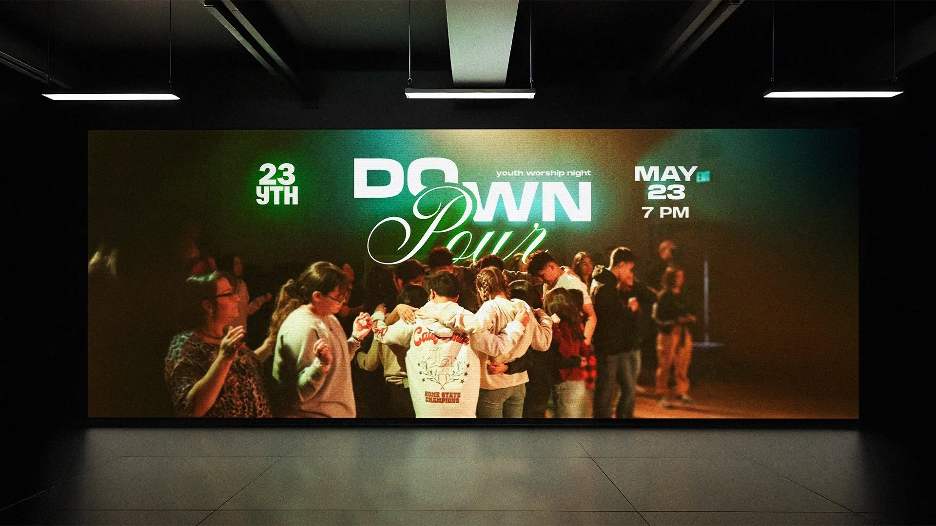 A group of young people gathered on stage for a youth worship night event, with a large screen behind them displaying the event details: date, time, and name, "Down Pour." The event is scheduled for May 23rd at 7 PM, with the words "youth worship night" visible on the screen.