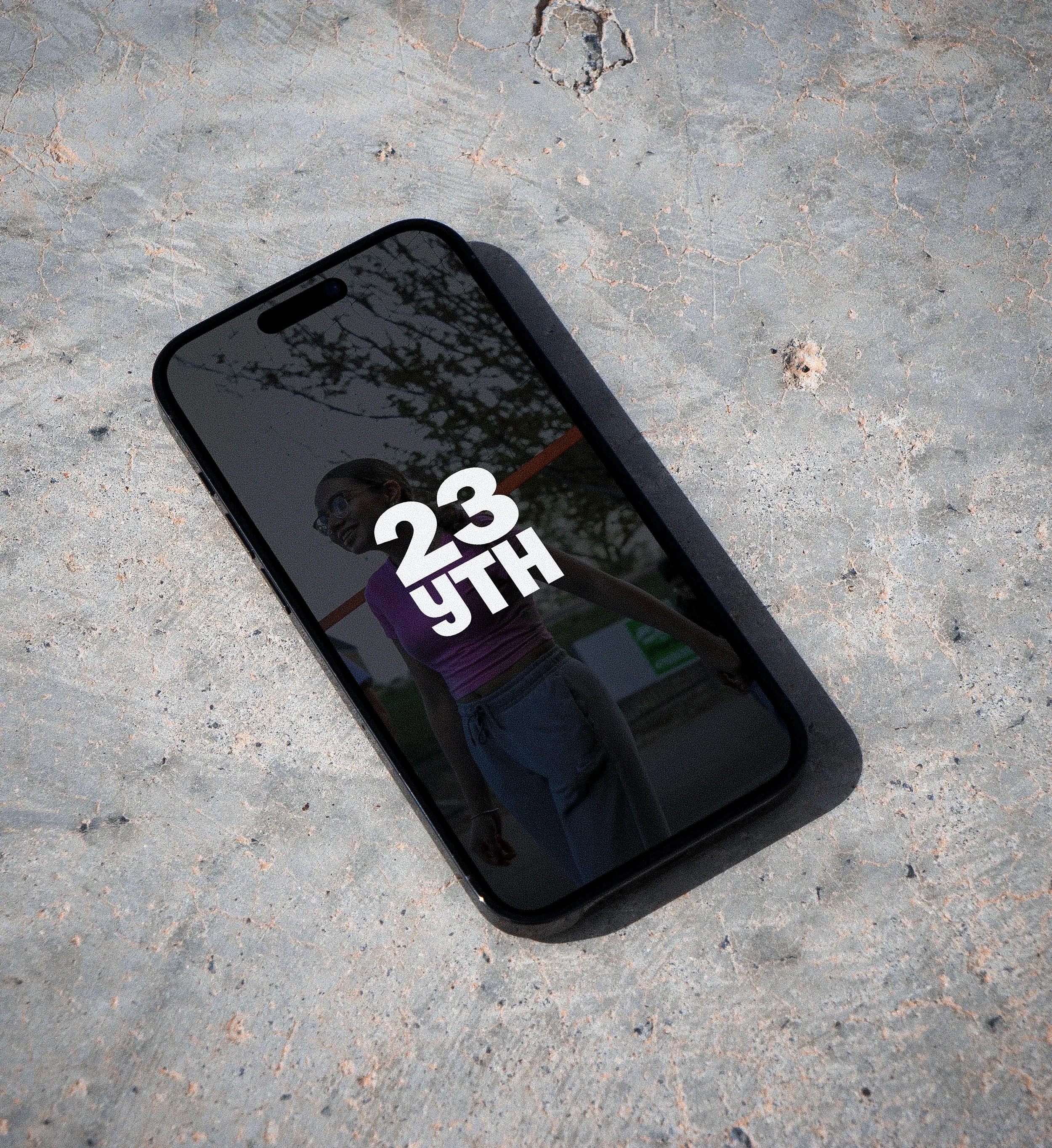 Close-up of a smartphone lying on a cracked concrete surface, displaying the time 23:48 and a reflection of trees and a person on its screen.