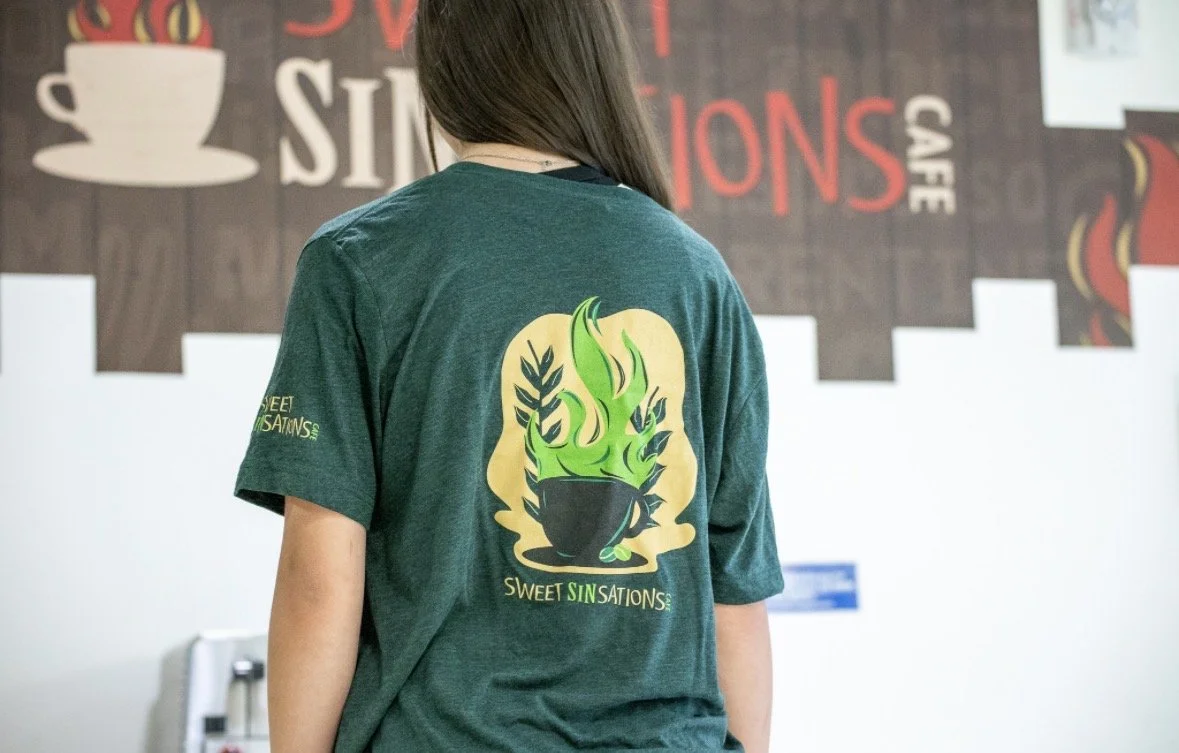 Back of a person wearing a green T-shirt with a graphic of a steaming cup and herbs, and the words "Sweet Sin Stations" on it, inside a cafe.