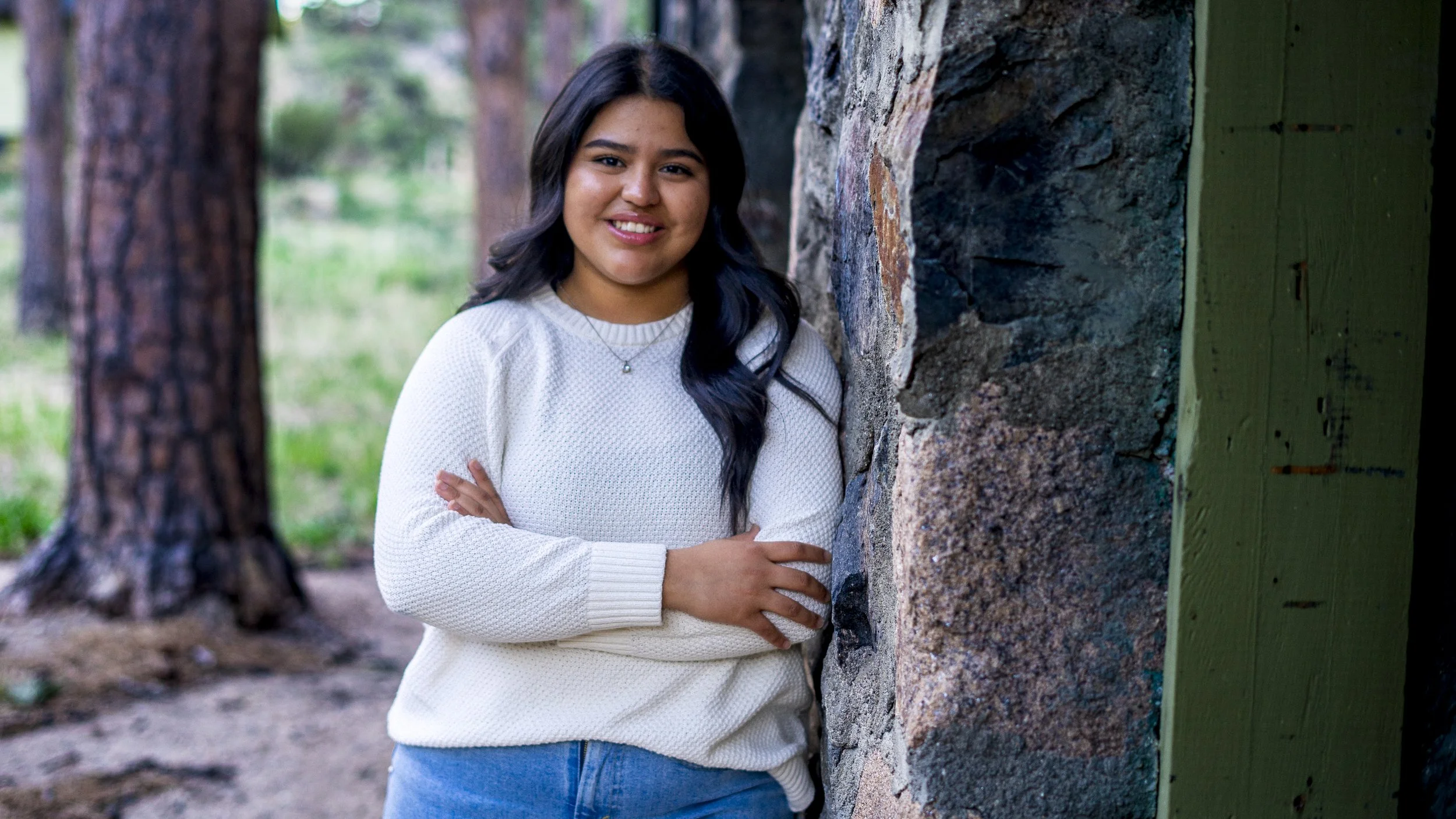 A young woman with dark hair wearing a white sweater and blue jeans, smiling and standing outdoors against a stone wall with trees in the background.