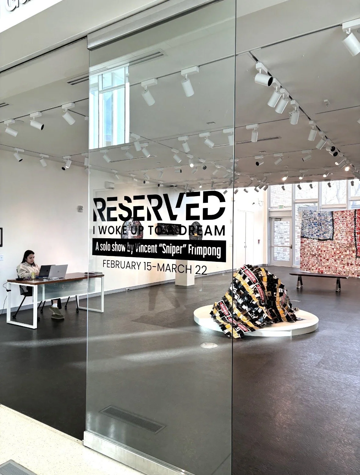 Interior of an art gallery with a promotional poster on the glass wall for an exhibition titled 'RESERVED I WOKE UP TO DREAM,' featuring solo show by Vincent 'Sniper' Frimpong running from February 15 to March 22. The gallery contains an art piece ma