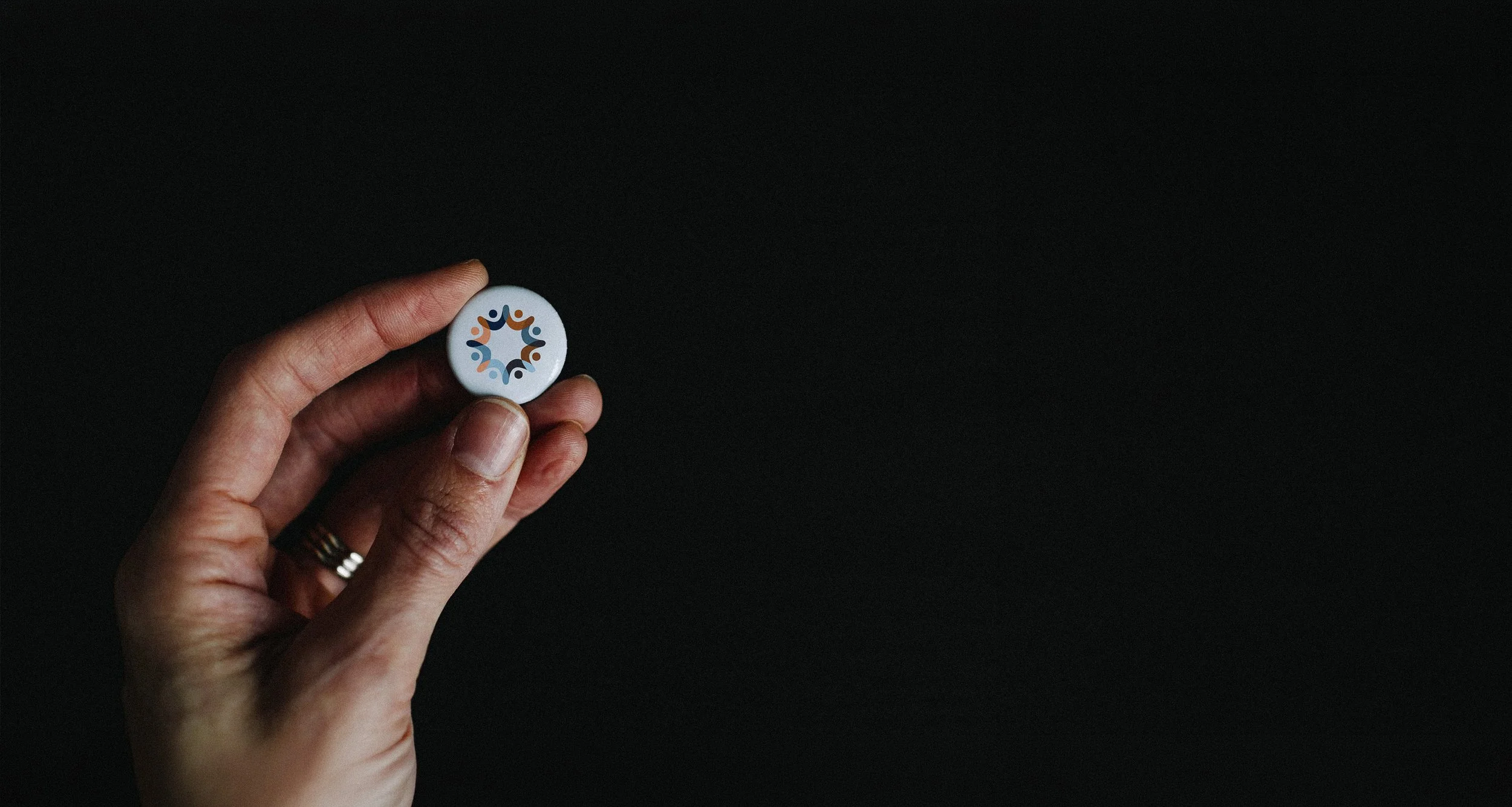 A person holding a circular badge with a colorful logo against a black background.