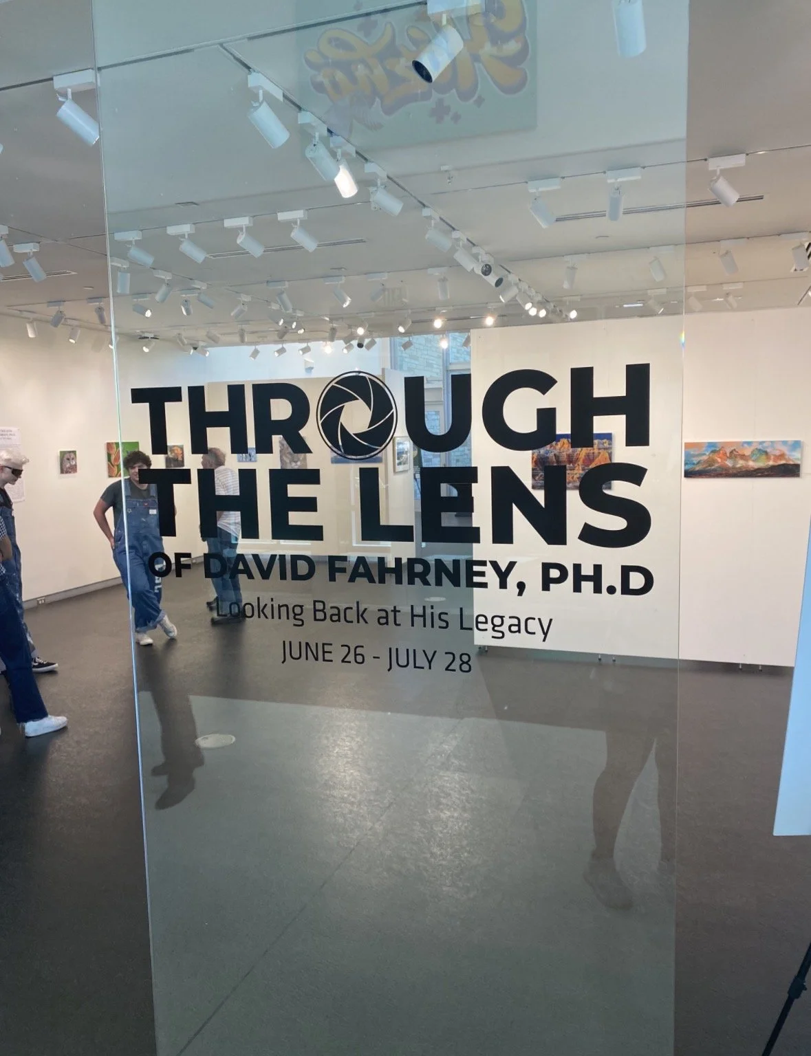 Exhibition announcement on glass door for 'Through the Lens of David Fahney, Ph.D., Looking Back at His Legacy,' with dates June 26 to July 28, and reflections of people and artwork inside the gallery.