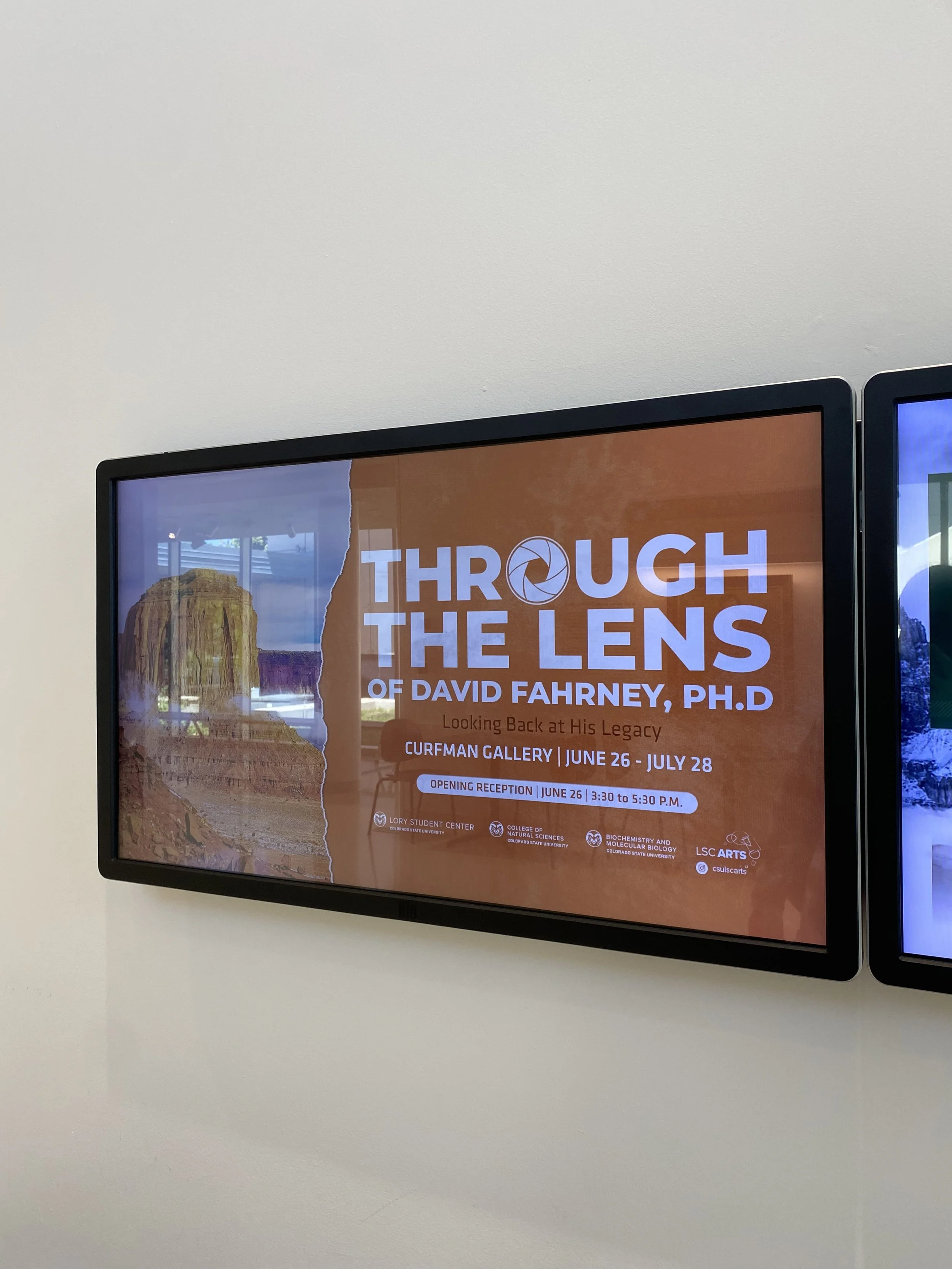 Digital display board with an exhibit invitation titled 'Through the Lens of David Fahrney, Ph.D.' at Curfman Gallery shows a background image of a desert landscape with rock formations.