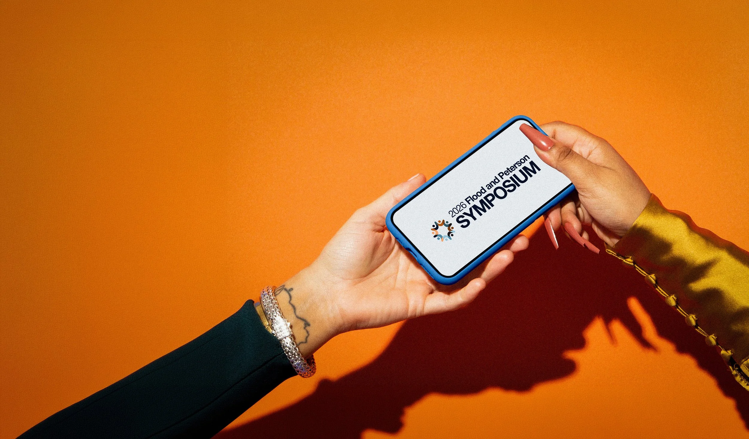 Person holding a smartphone displaying text for the 2026 Flood and Peterson Symposium against an orange background with their shadow cast on the wall.