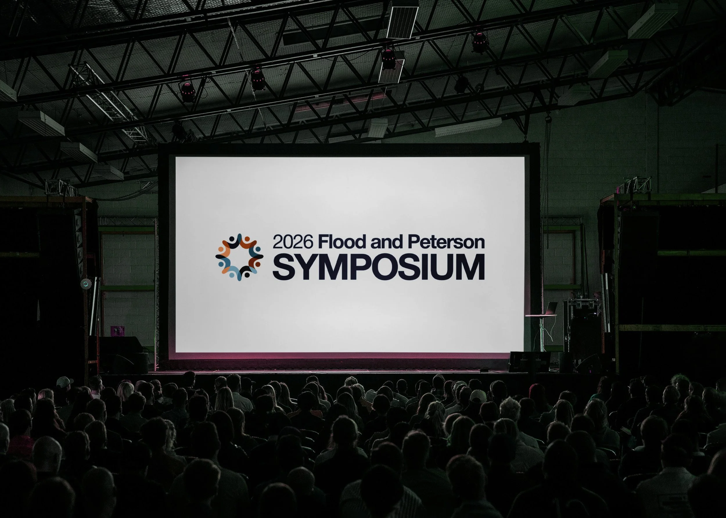 Audience attending the 2026 Flood and Peterson Symposium in a large indoor venue with a stage featuring a large illuminated screen displaying the event name and logo.