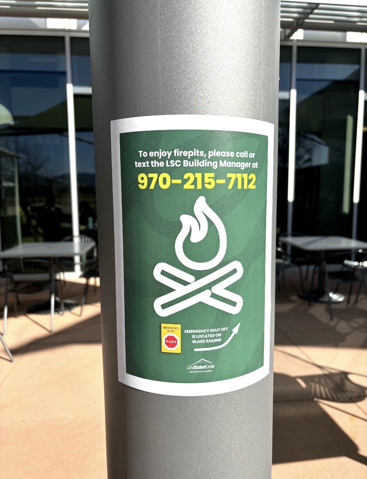 A green sign on a pole with a firepit icon, instructions to call or text a phone number to enjoy firepits, and a push button for emergency stop, located on a glass railing outside a building.