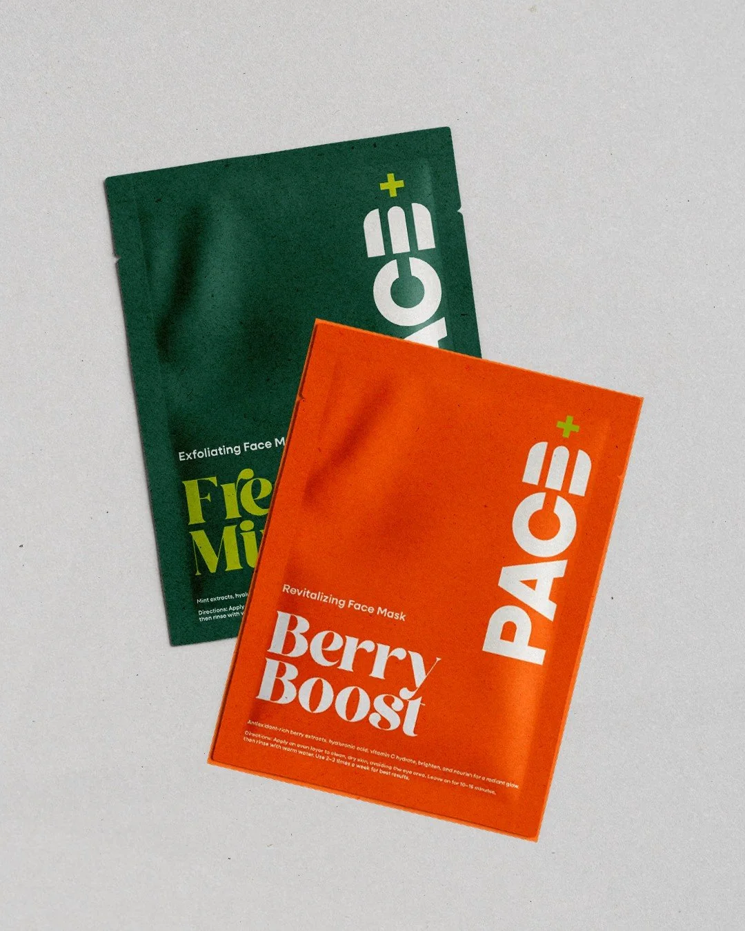 Two colorful face mask packets, one orange labeled Berry Boost and one green labeled Fresh Mint, lying on a white surface.