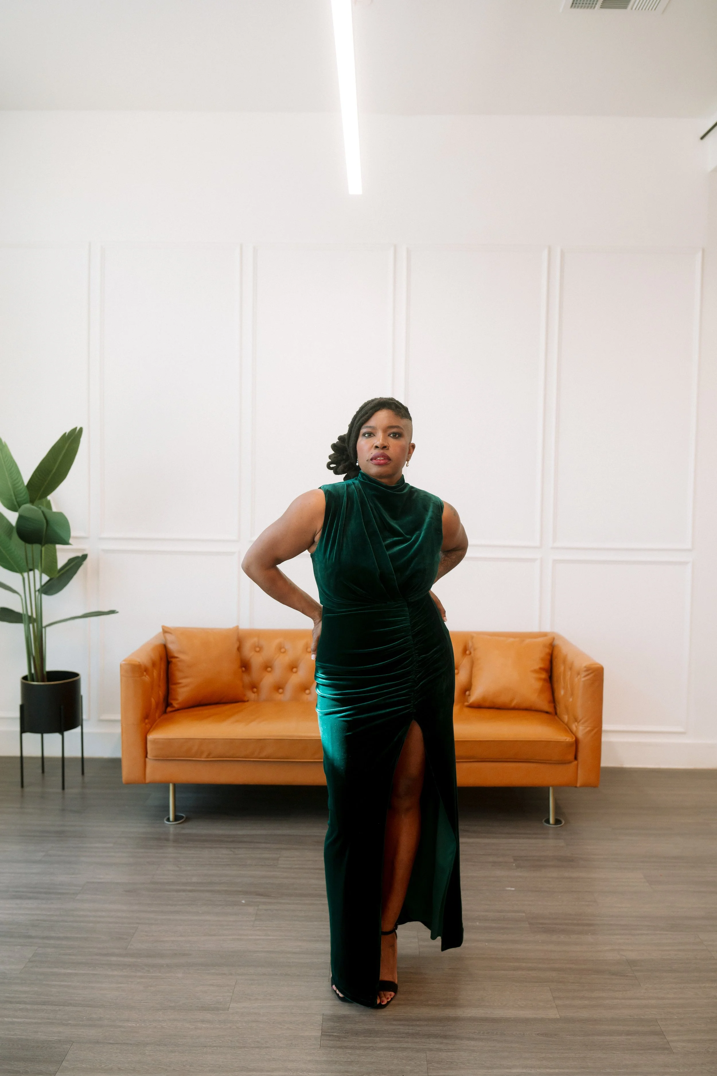 A woman in a dark green velvet dress with a thigh-high slit standing in front of a tan sofa in a modern, minimalist room with white paneled walls, a tall green potted plant, and a wooden floor.