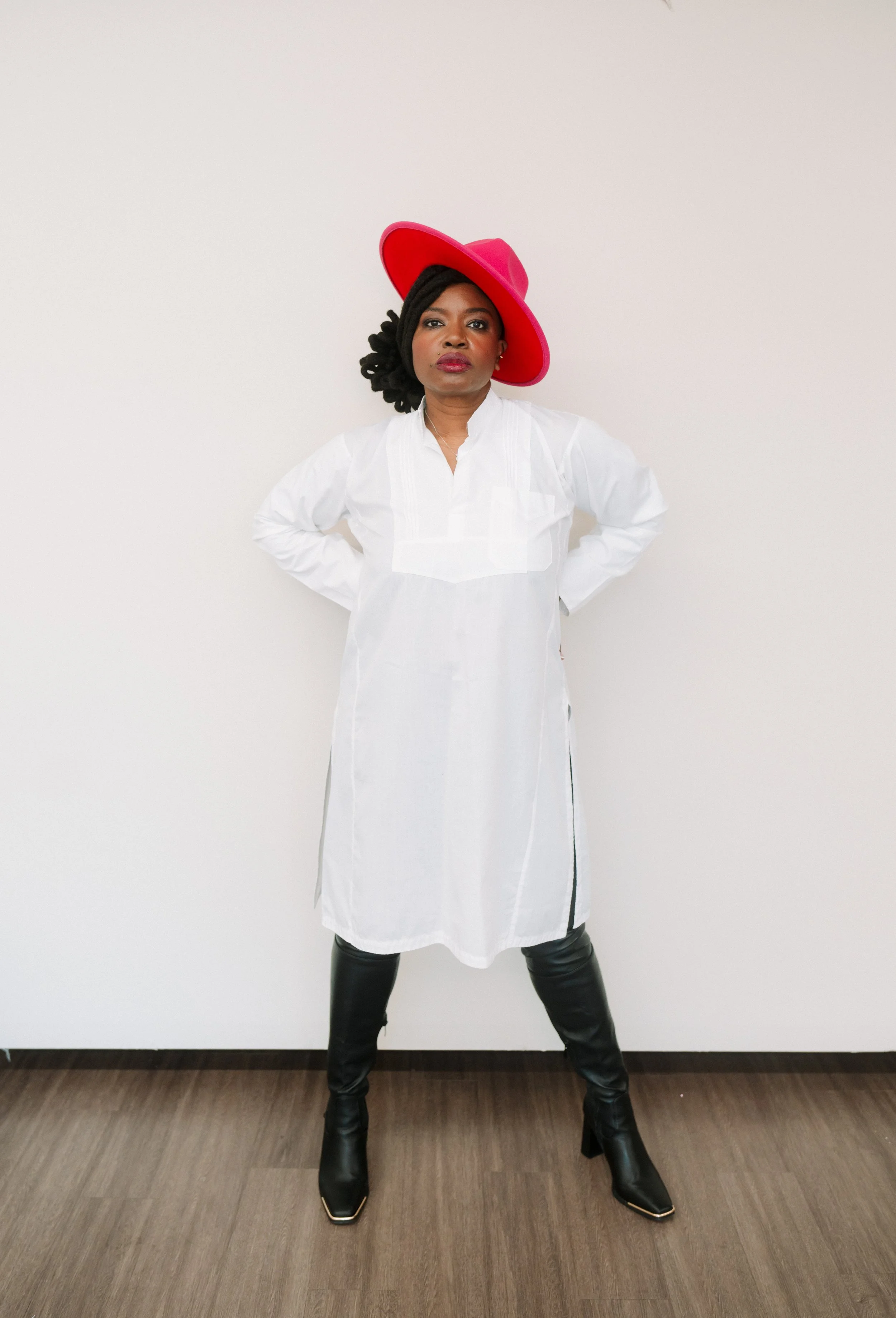 A woman wearing a large red hat, black boots, a white dress, and black leggings standing against a plain white wall with her hands on her hips.