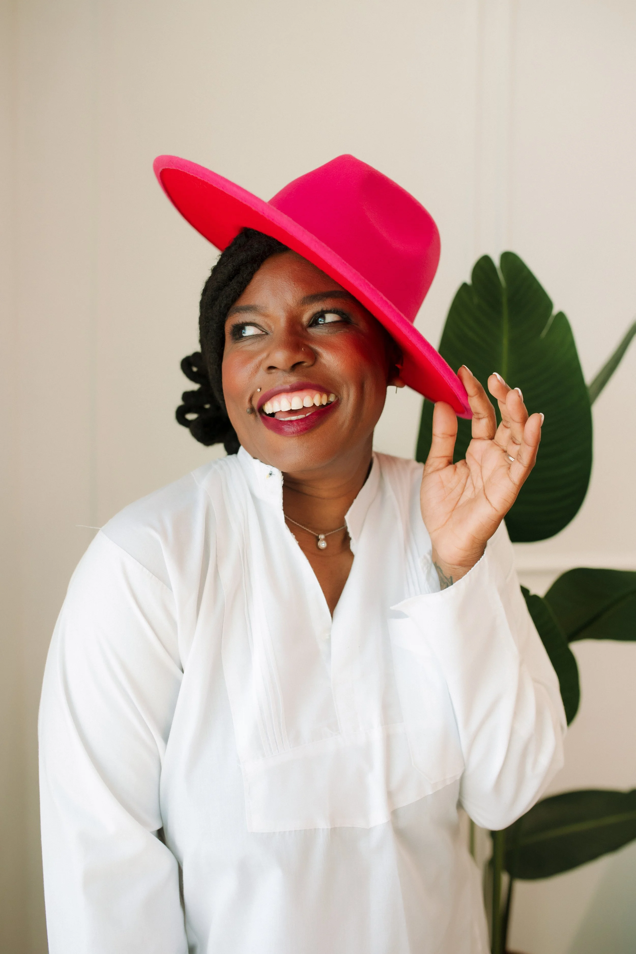 A woman with dark braided hair wearing a white top and a pink wide-brimmed hat is smiling and holding the brim of her hat. She is standing indoors with green plant leaves in the background.