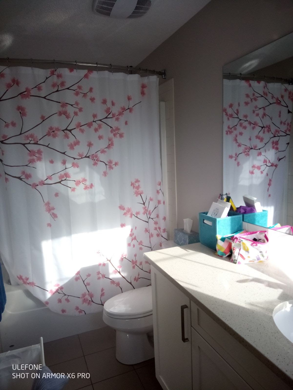 A bathroom with a white tub and a floral shower curtain featuring pink cherry blossoms on dark branches. There is a white toilet and a white sink cabinet with a countertop holding a blue organizer with various items, and a tissue box. A mirror reflects part of the shower curtain and wall.