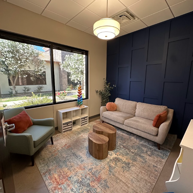 A cozy living room with a beige sofa and two throw pillows, a green armchair with an orange pillow, a white shelf, and two small wooden stools. Large window showing trees and outdoor greenery. Blue accent wall, ceiling light fixture, patterned rug.