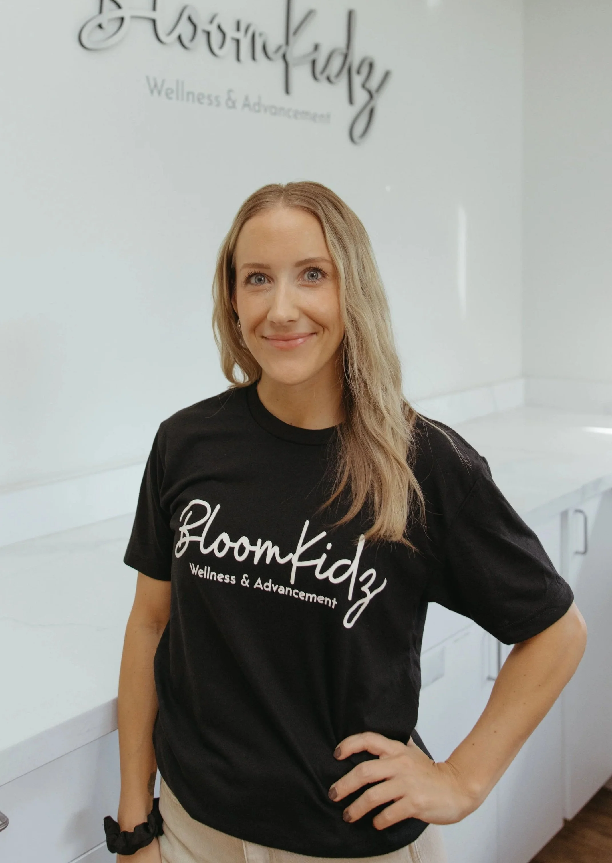 A woman with long, wavy blonde hair, wearing a black t-shirt with white text that reads 'BloomKidz Wellness & Advancement,' standing in front of a white wall with a sign that says the same, She is smiling and has her right hand on her hip.