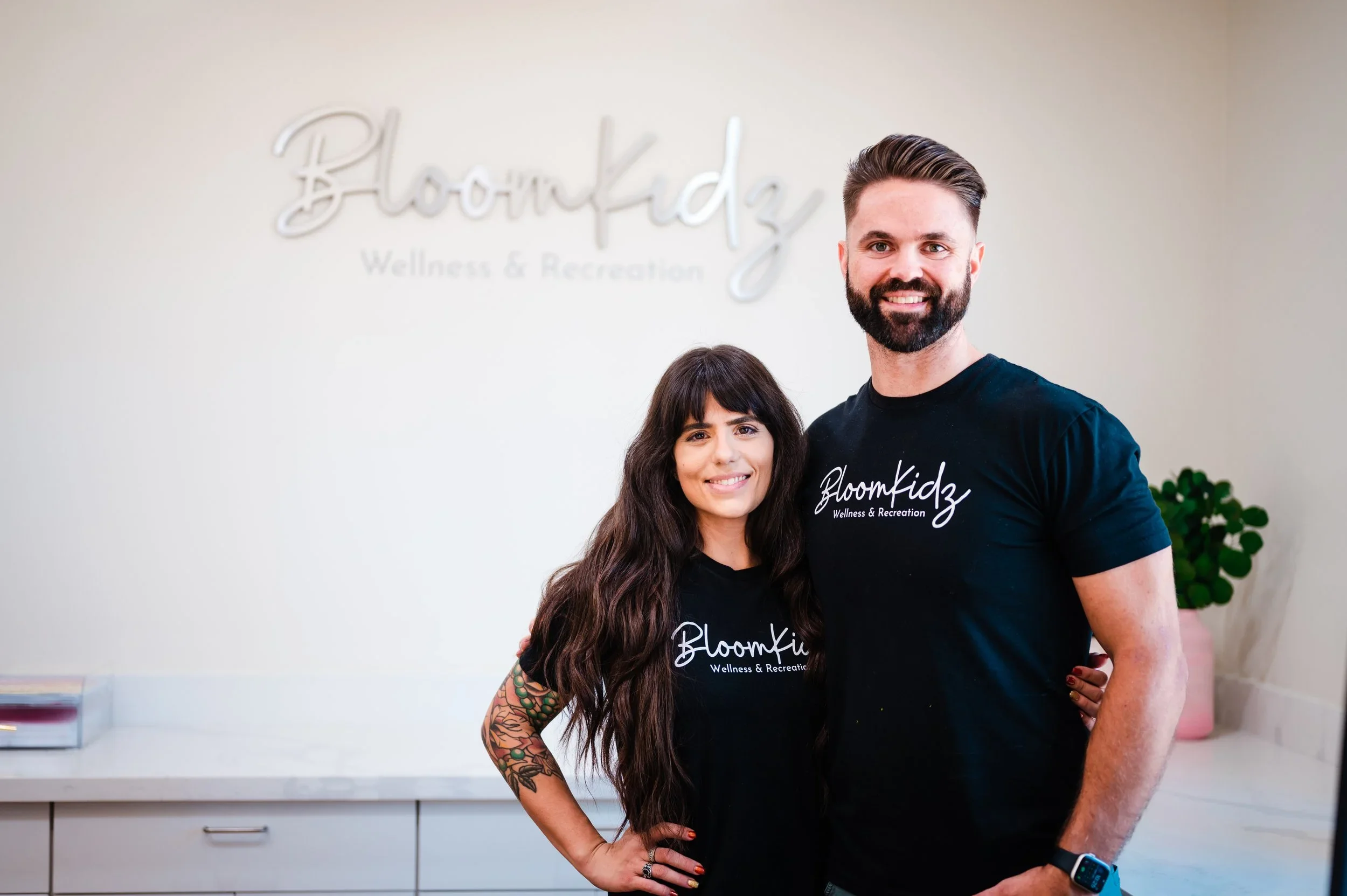 A man and a woman standing together in front of a wall with a sign that reads 'Bloomkidz Wellness & Recreation'. Both are smiling, wearing matching black t-shirts with the same logo, and the woman has tattoos on her arm.