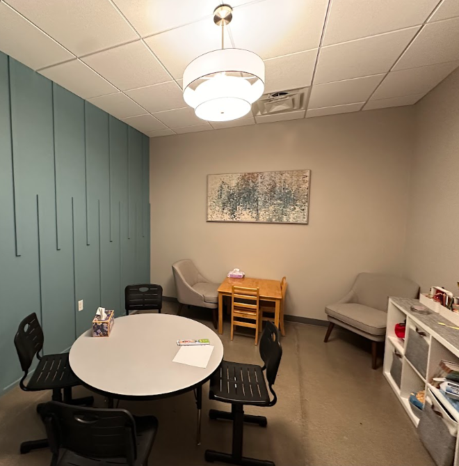 A small meeting room with a round table, four black chairs, a beige armchair, a gray loveseat, a white bookshelf, and a small wooden table with two chairs. There's a large abstract painting on the beige wall, a modern ceiling light, and teal paneling