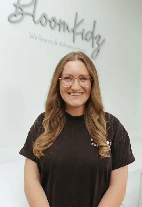 A smiling woman with long wavy hair and glasses standing in front of a wall with a sign that reads "BloomKidz Wellness & Advancement."