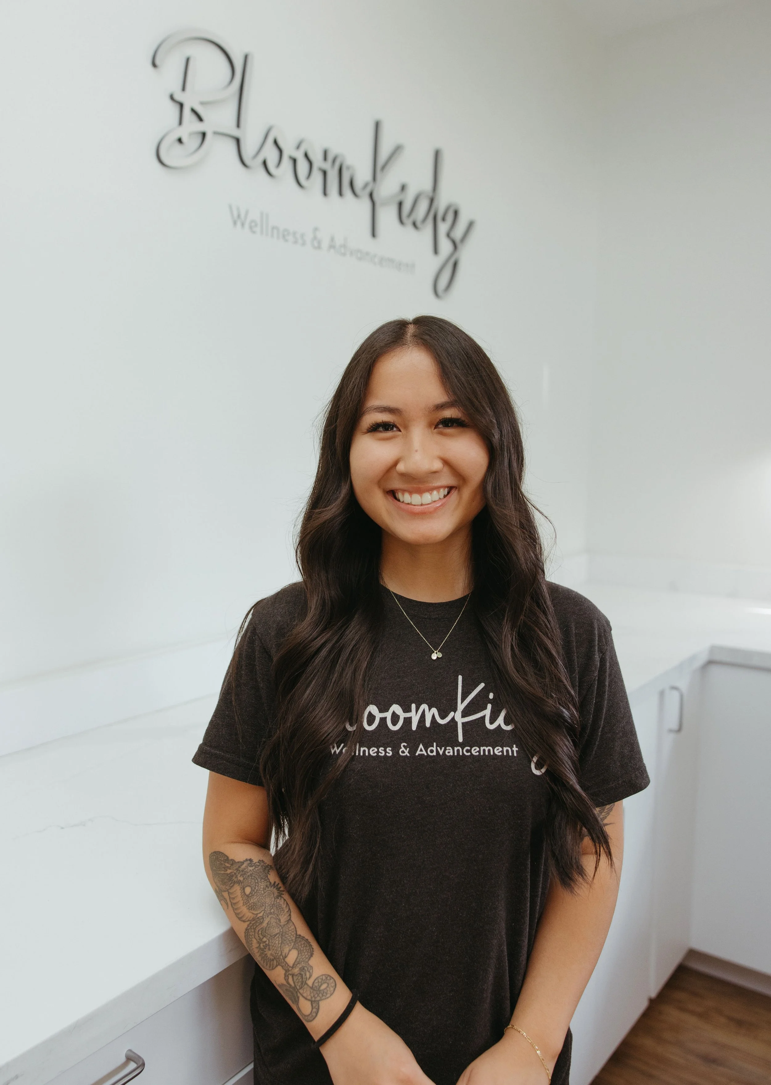 A woman with long dark hair smiling, standing in front of a white wall that reads 'BloomKidz Wellness & Advancement'. She is wearing a black t-shirt with the same text, and has a tattoo of flowers and a snake on her left arm.