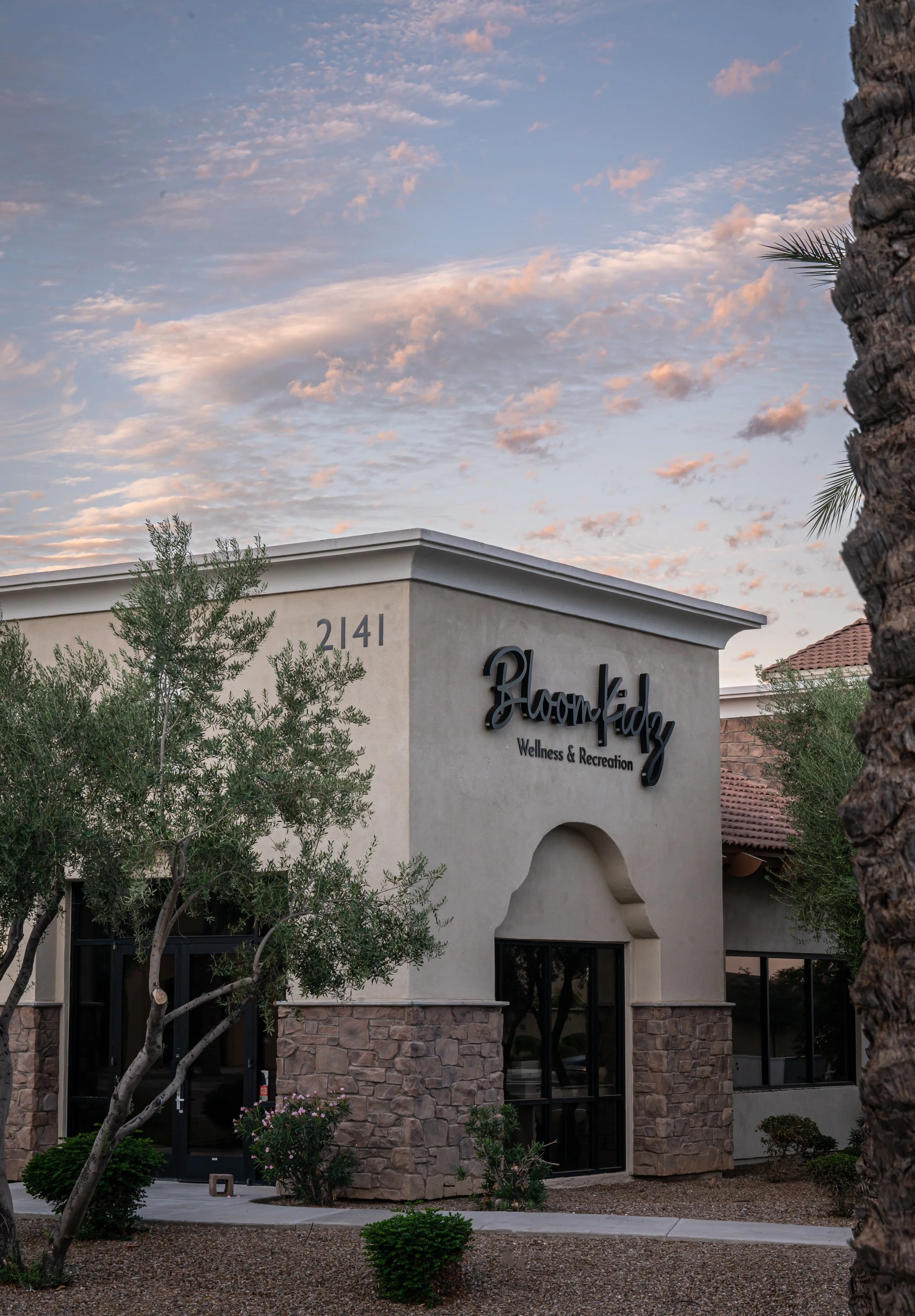 Exterior of Bloomify Wellness & Recreation building at sunset, with stone walls, landscaping, palm trees, and cloud-filled sky.
