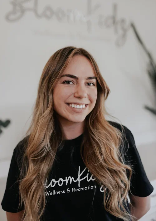 Young woman with long wavy blond hair smiling, wearing a black t-shirt with the words 'BloomKidz Wellness & Recreation' on it, standing in front of a light-colored wall with some decor.