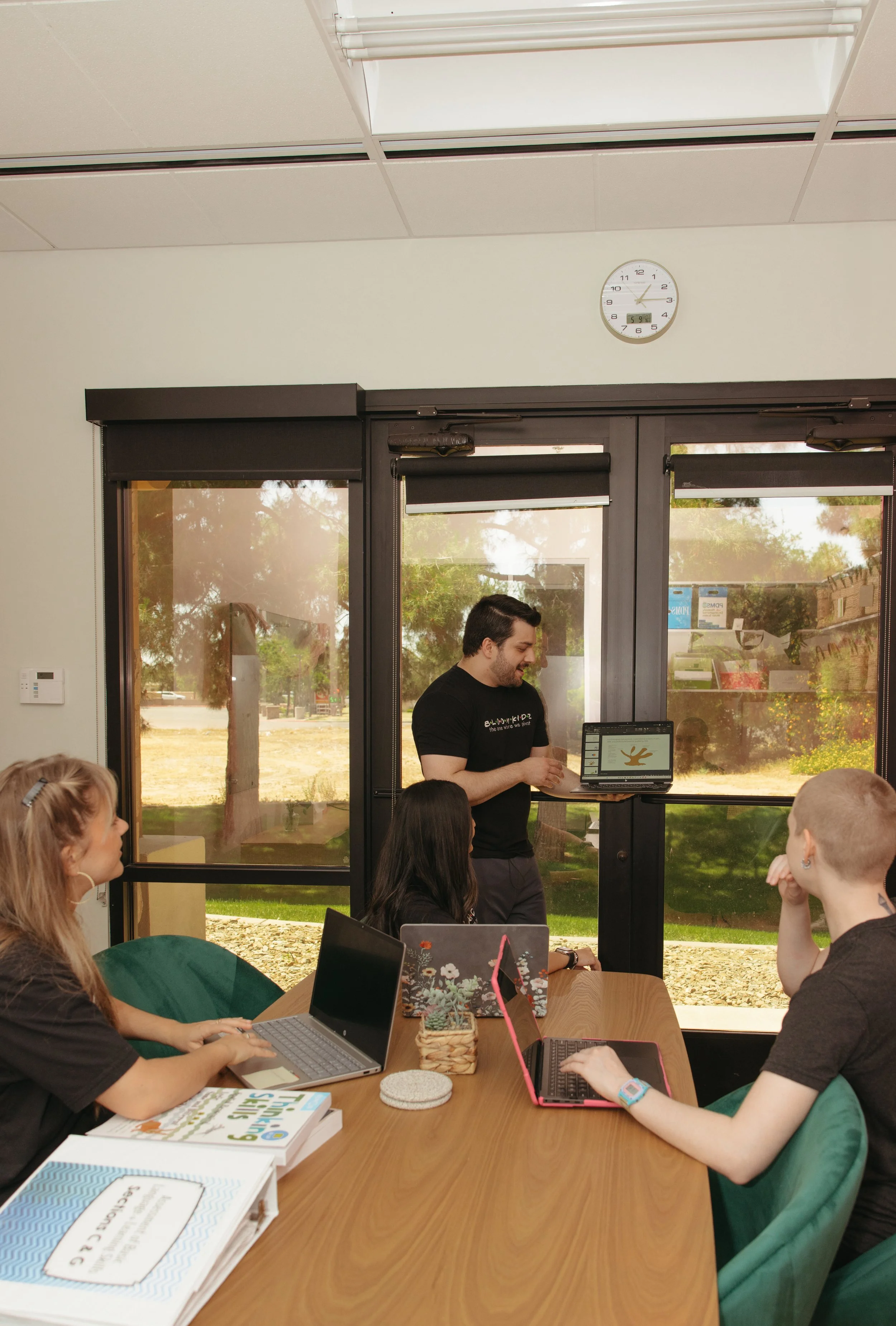 A man standing in front of a group of people, giving a presentation in a room with large glass windows showing an outdoor view. The group has laptops and books on the table