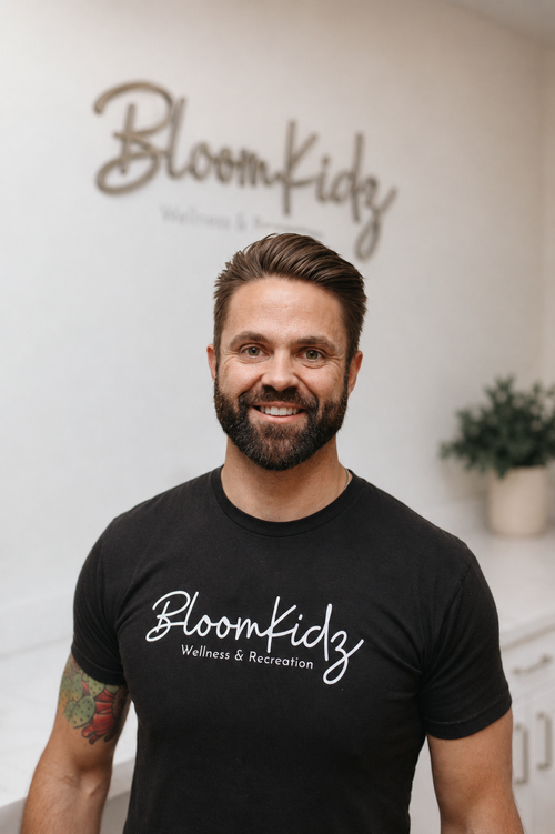 Man with beard and tattoo wearing a black Bloomkidz Wellness & Recreation t-shirt, smiling in front of a Bloomkidz sign and potted plant.