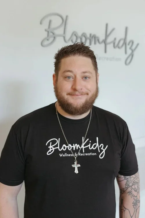 A man with a beard and short hair standing indoors, wearing a black t-shirt with 'BloomKidz Wellness & Recreation' written on it, a silver necklace with a cross, and sporting tattoos on his left arm.