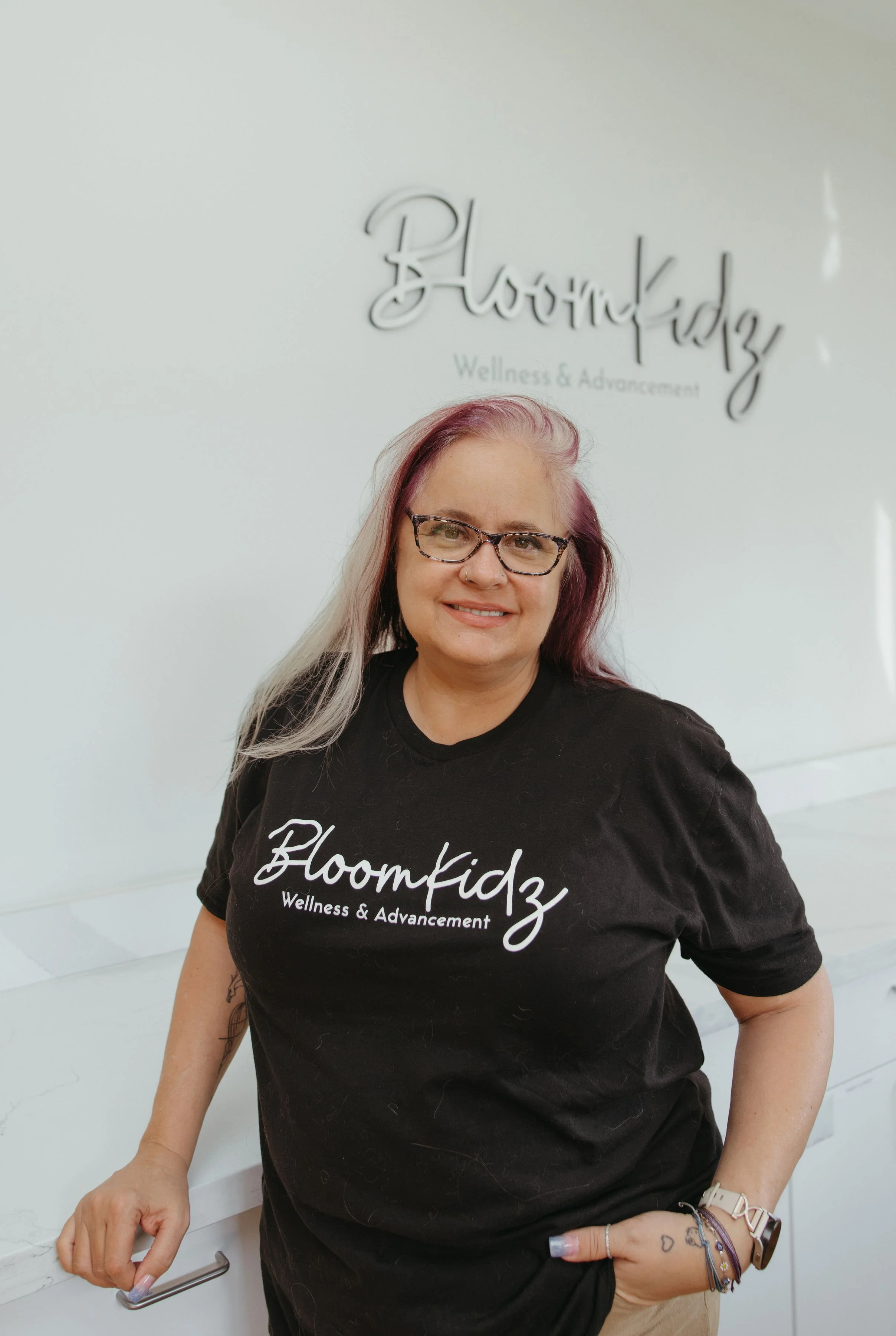 Smiling woman with glasses and dyed hair, wearing a black T-shirt that says 'Bloomkidz Wellness & Advancement,' standing in front of a light-colored wall with a sign that reads 'Bloomkidz Wellness & Advancement.'