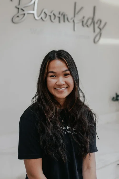 A smiling young woman with long dark hair standing inside a modern, well-lit room with a white background and a sign that says "BloomKidz" in cursive above her head.
