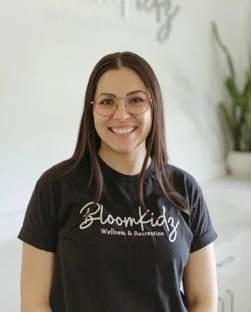 A smiling woman wearing glasses and a black t-shirt with the text 'BloomKidz Wellness & Recreation' standing indoors with a white wall and a potted plant in the background.