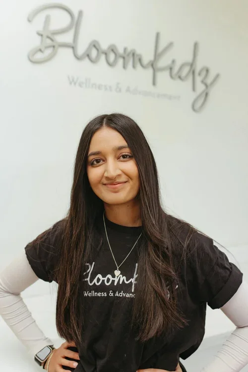 A woman with long dark hair smiling, wearing a black t-shirt and white long sleeves underneath, standing in front of a wall with the 'BloomKidz Wellness & Advancement' logo.