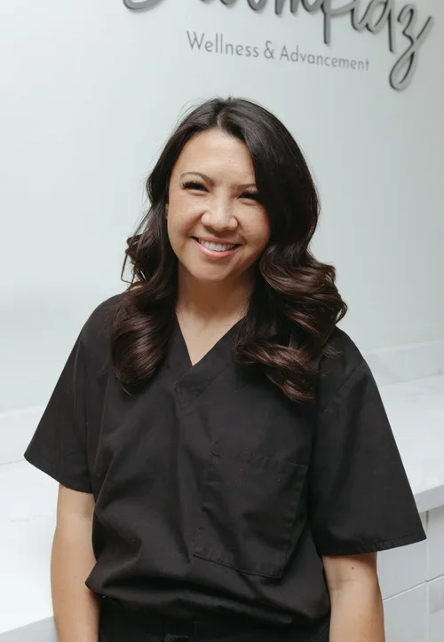 A woman with long wavy dark hair, wearing a black medical uniform, smiling at the camera in a wellness center or clinic.