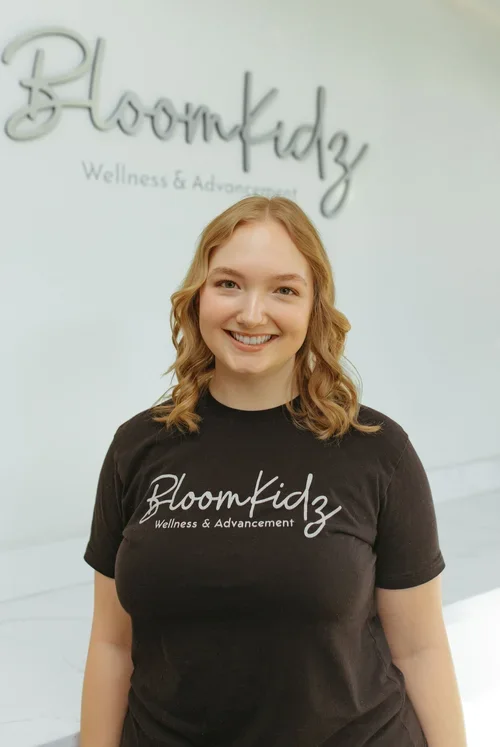 A young woman with blonde, wavy hair smiling while wearing a black t-shirt with the BloomKidz Wellness & Advancement logo, standing in front of a wall with similar logo text.