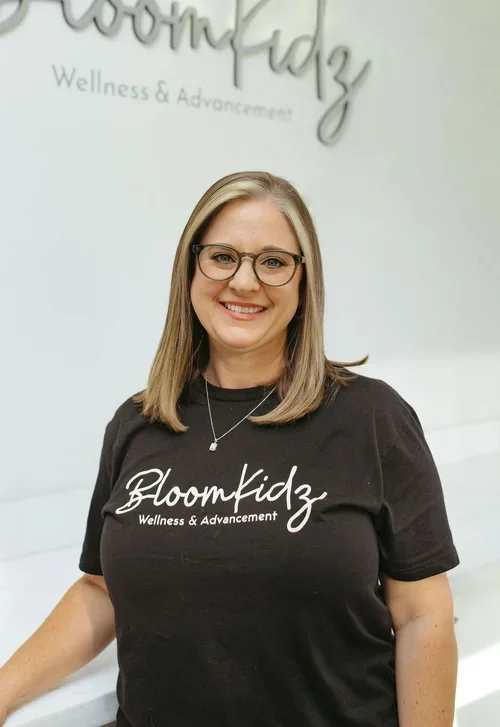 Woman with glasses and blonde hair wearing a black BloomKidz Wellness & Advancement t-shirt, smiling in a wellness center.