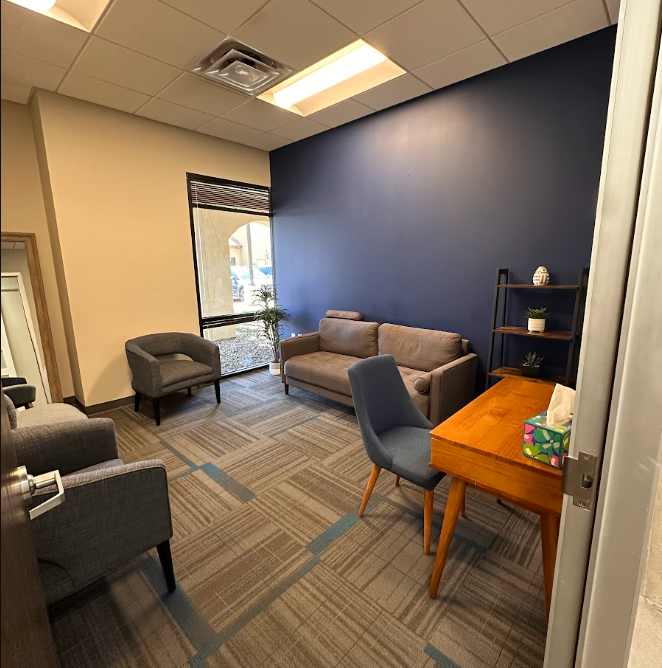 A small waiting or office area with a beige couch, gray armchair, and a wooden desk with a blue chair. There is a black shelf with decorative items and a tissue box on the desk. A large window lets in natural light, and a potted plant is near the win