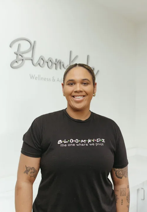 A smiling woman with dark hair tied back, wearing a black Bloomkidz t-shirt, standing in front of a wall with Bloomkidz Wellness & Advancement logo.