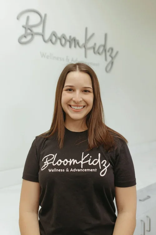Smiling woman wearing a BloomKidz Wellness & Advancement T-shirt in front of a BloomKidz logo wall