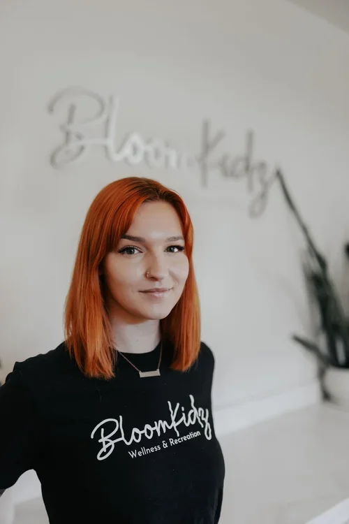 A young woman with shoulder-length red hair, wearing a black shirt with the words 'Bloom Kidz Wellness & Recreation,' standing indoors with a blurred background of a white wall and a plant.