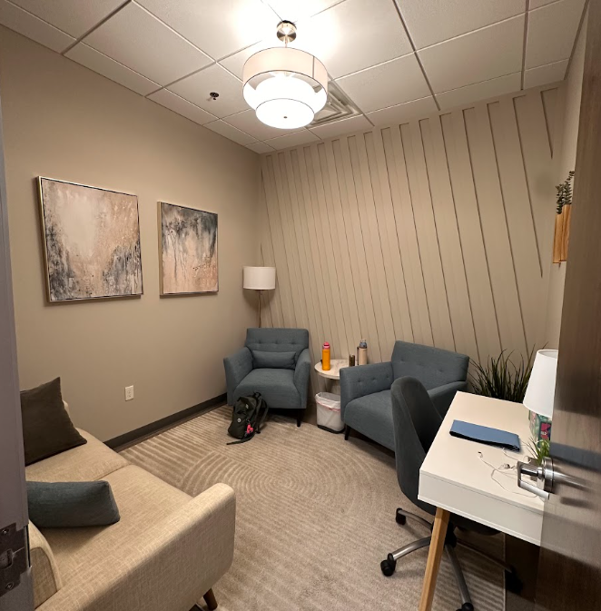 A small, cozy room with beige walls and a beige carpet. Contains a beige sofa with brown and beige pillows, two blue armchairs, a white desk with a black chair, a small side table with bottles and snacks, a white lamp, and wall art with abstract desi