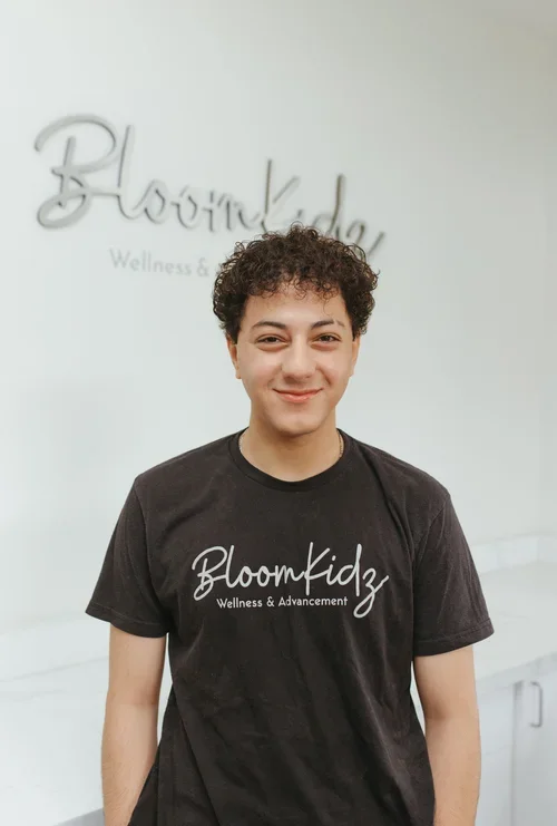 Young man smiling, wearing a black T-shirt that says 'Bloomkidz Wellness & Advancement,' standing in front of a white wall with the 'Bloomkidz Wellness & Advancement' logo.