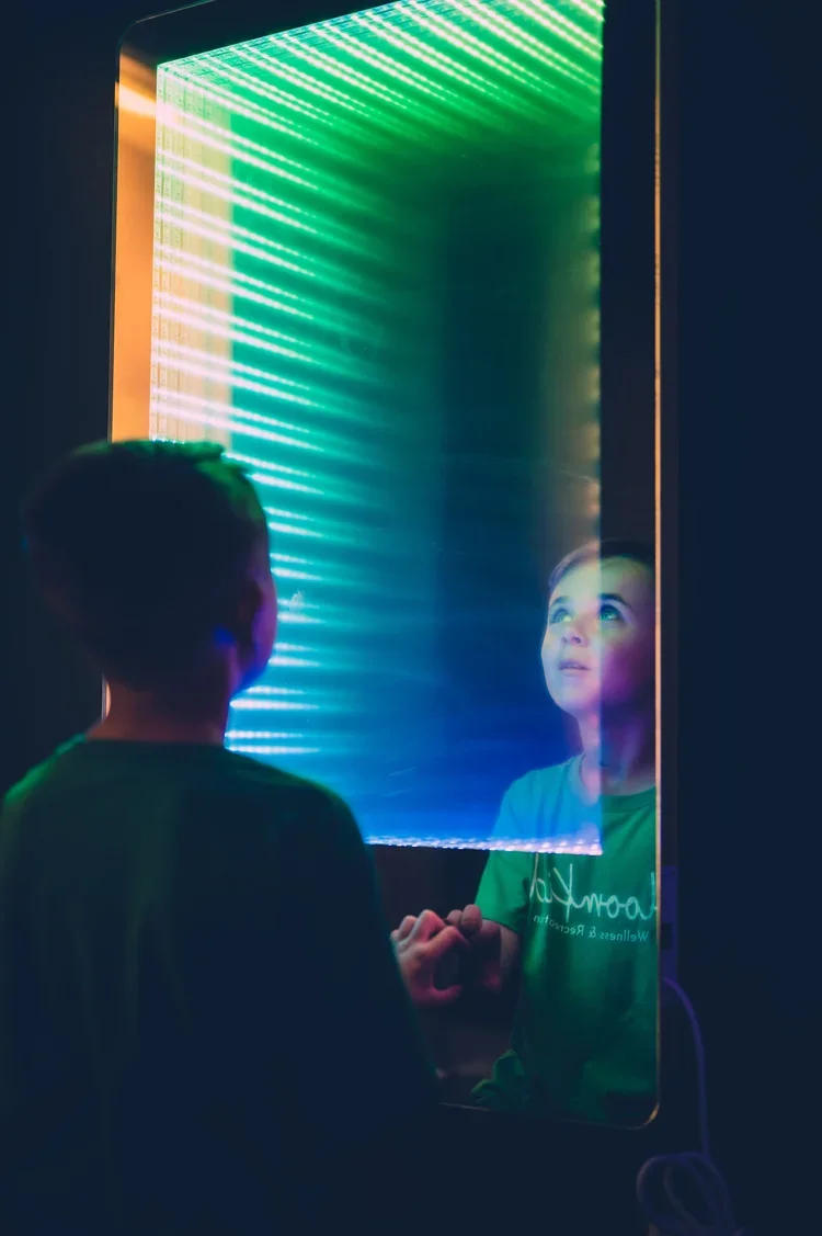 A boy standing in front of a glass display with colorful, animated LED lights, looking at his reflection.