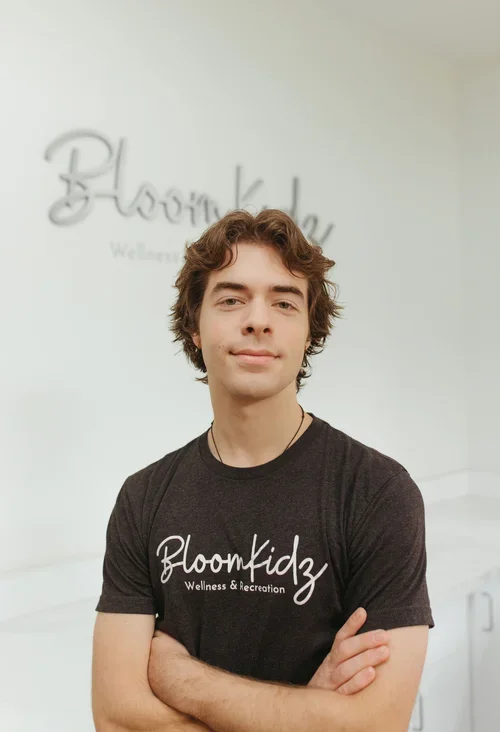 Young man with curly hair wearing a BloomKidz Wellness & Recreation T-shirt, standing with arms crossed in front of a wall with a BloomKidz sign.