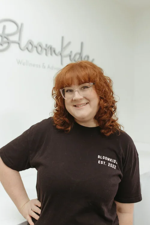 A woman with curly red hair wearing glasses and a black T-shirt that says Bloomkidz Est. 2022, standing indoors with a sign that reads Bloomkidz Wellness & Advancement in the background.