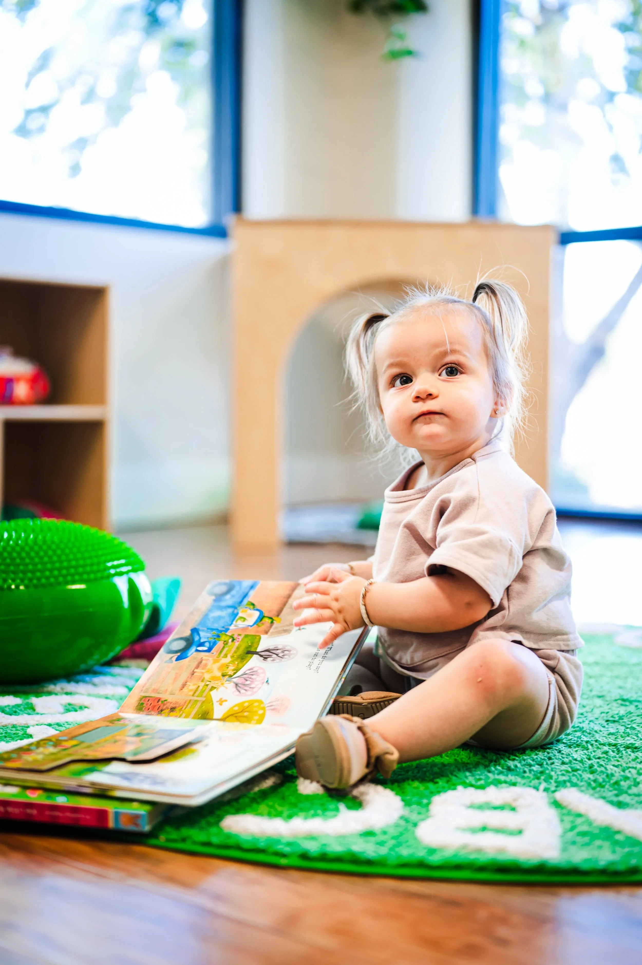 A young girl with blonde hair in pigtails, sitting on a green and white patterned rug indoors, holding a colorful children's book. In the background, there are large windows with a view of trees and a wooden playhouse.